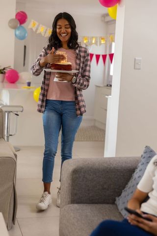 African american woman carrying birthday cake into living room for surprise celebration