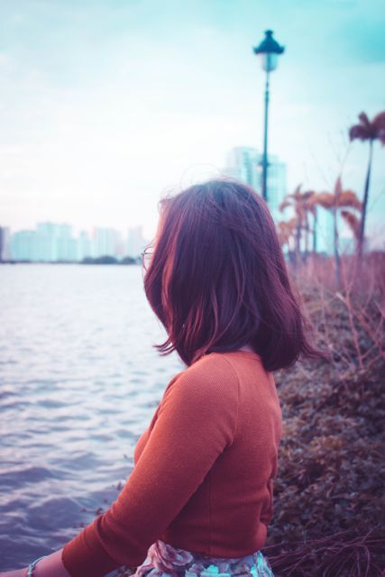 Woman sitting near the water with an urban skyline in the background. Suitable for concepts of contemplation, relaxation, urban outdoor lifestyle, and calming moments. Can be used in travel brochures, lifestyle blogs, or mental wellness articles.