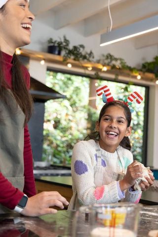 Diverse mother and daughter kneading dough laughing during holiday baking in bright kitchen
