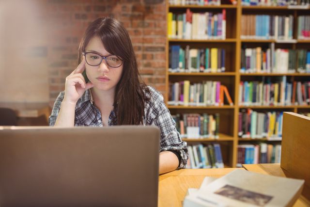 Young woman with glasses using laptop in library, appearing thoughtful. Ideal for educational content, online learning platforms, academic research, and technology in education themes.