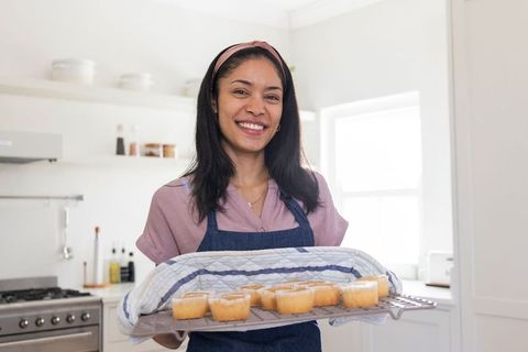 Home baker smiling while holding freshly baked cupcakes on cooling rack in sunlit kitchen