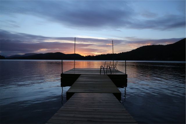 Wooden dock extending into calm lake with beautiful reflective water at sunset. Mountains form a dark silhouette in background, adding to peaceful environment. Ideal for illustrating themes of nature, travel, relaxation, meditation, and tranquility.