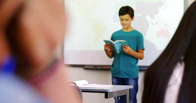 Boy wearing green shirt reading book in front of whiteboard with world map. Ideal for content on education, classroom activities, improving public speaking skills, and young learners developing confidence.
