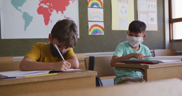 Happy diverse boys in facemasks sitting at desks and writing. School, learning, childhood, education, unaltered, health, hygiene, coronavirus.