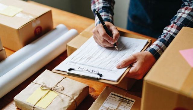 Checking shipping checklist at packing table with parcels, clipboard, kraft paper packages