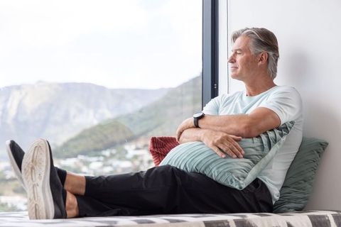 Senior man relaxing on window seat hugging pillow, gazing at panoramic mountain view