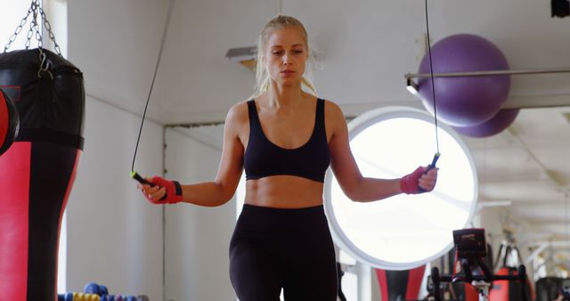 A young Caucasian woman is focused on her workout, skipping rope in a gym environment, with copy space. Her athletic attire and the equipment around her underscore a commitment to fitness and health.