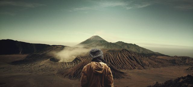 Person dressed in winter clothing facing rugged mountains with an active volcano in the distance during sunrise. Ideal for travel blogs, adventure tourism websites, and motivational content about exploration and connecting with nature.
