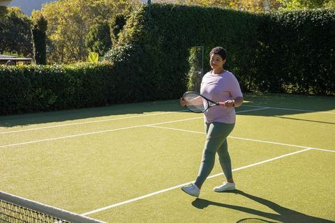 African american woman walking on grass tennis court holding racket under sunny sky