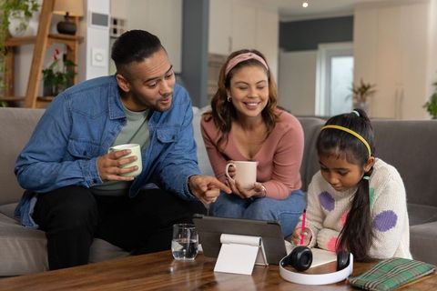 Parents guiding daughter with tablet and notebook on living room couch, cozy study moment