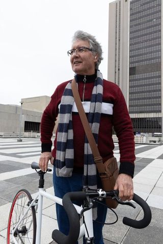 Senior commuter holding road bicycle on urban plaza wearing striped scarf and satchel
