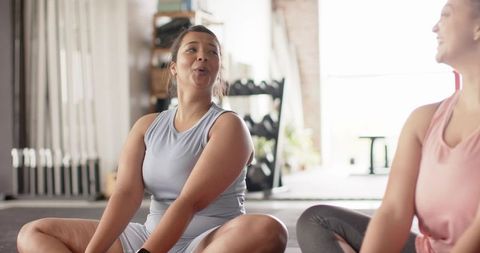 Two women stretching and laughing on mats in bright gym with weights and natural light
