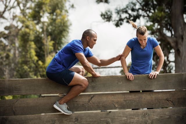 Trainer assisting woman climbing wooden wall during outdoor obstacle course. Ideal for use in fitness, teamwork, and training-related content. Perfect for illustrating concepts of support, motivation, and physical challenges.