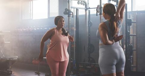 Diverse women stretching and chatting in gym near squat rack and free weights pre-workout