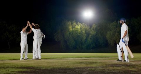 Night cricket match under floodlights featuring diverse players celebrating wicket