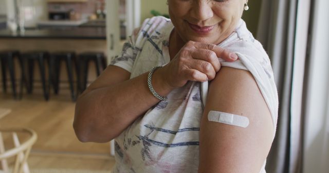 Smiling woman showing her bandaged arm inside home environment after getting vaccinated. Ideal for use in healthcare campaigns, immunization awareness, or medical service advertisements.