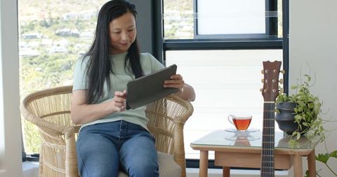 Asian woman sitting in rattan chair using tablet with tea and guitar by sunlit window