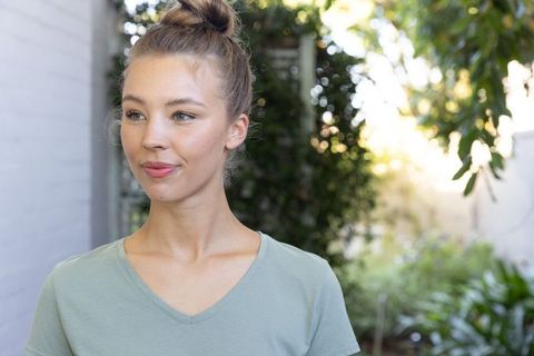 Young woman standing in sunlit backyard looking left wearing green v-neck and top knot