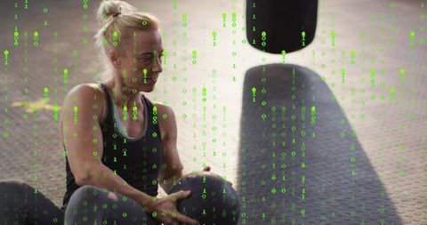 Fit woman sitting holding medicine ball on gym mat with green binary data overlay