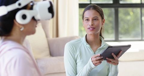 Diverse women testing vr headset in living room with instructor guiding using tablet