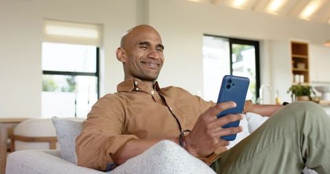 African american man relaxing on couch and scrolling phone in bright modern living room
