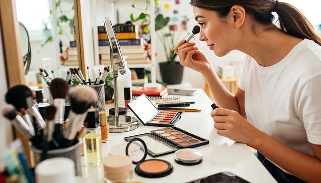 Young woman applying cheek makeup at bright home vanity with brushes and palettes, mirror