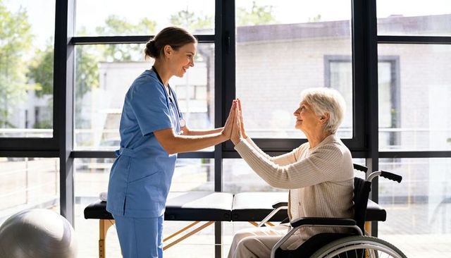 Nurse supporting senior in wheelchair giving high-five during physical rehabilitation