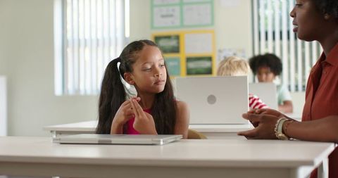 Young student adjusting fingers at classroom desk with laptop while teacher guiding