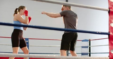 Female boxer sparring with male trainer in boxing ring, red hand wraps, coaching workout