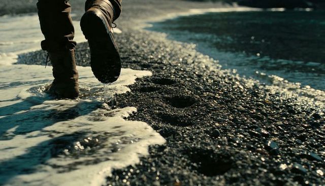 Walking boots leaving footprints on dark pebble beach with foamy surf and low-angle motion