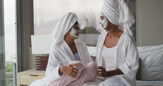 Image of relaxed diverse female friends moisturizing with face masks and talking in bathroom. Friendship and taking care of yourself and beauty concept.