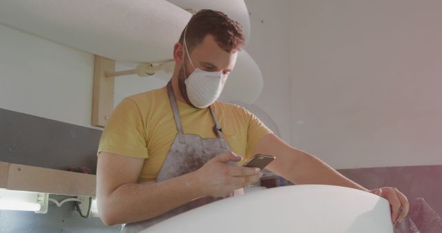 Craftsman inside workshop, wearing safety mask and apron, focused on checking smartphone. Background shows specialized workbench. Suitable for themes of crafts, safety in the workplace, use of technology in manual labor, and woodworking industry.