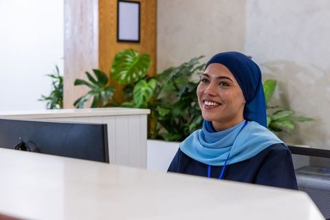 Smiling african receptionist in blue headscarf at modern office front desk with plants