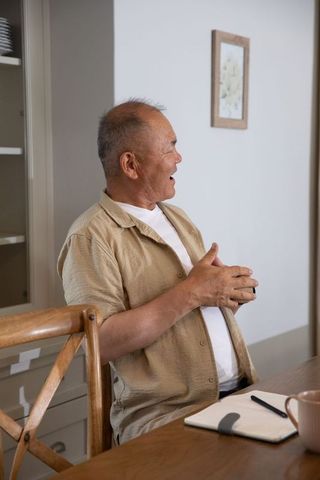 Senior chinese man gesturing and talking at cozy dining table with notebook and mug