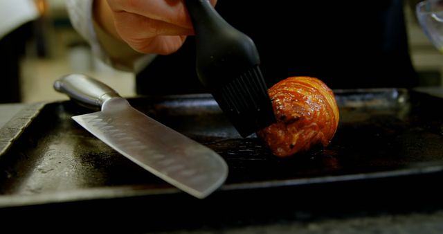 Chef glazing small piece of roasted meat on baking tray, sharp knife positioned nearby. Suitable for content related to culinary arts, gourmet cooking, kitchen skills, recipe articles, and food preparation. Ideal for websites, blogs, and social media posts focused on cooking and gastronomy.