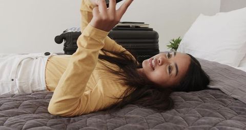 Asian woman lying on bed using smartphone with suitcase nearby for relaxed travel moment