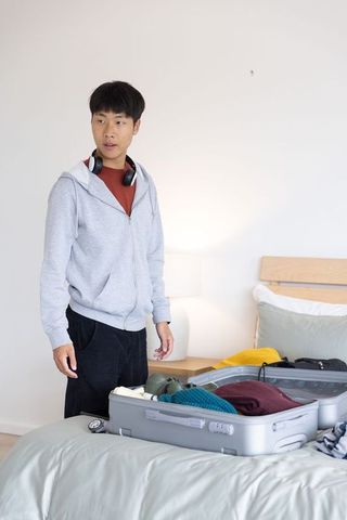 Asian man packing suitcase on bed with headphones preparing for weekend getaway