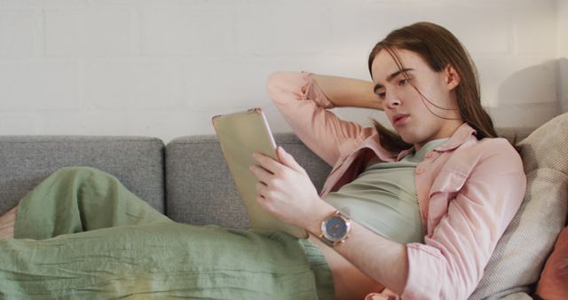 Young woman lying on couch reading tablet at home. Useful for depicting relaxation, leisure time, digital devices, at-home study, online entertainment and casual lifestyle contexts.