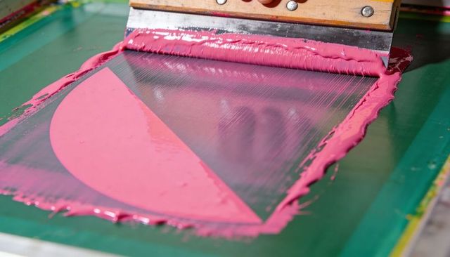 Screen printing process pulling bright pink ink with wooden squeegee over mesh stencil