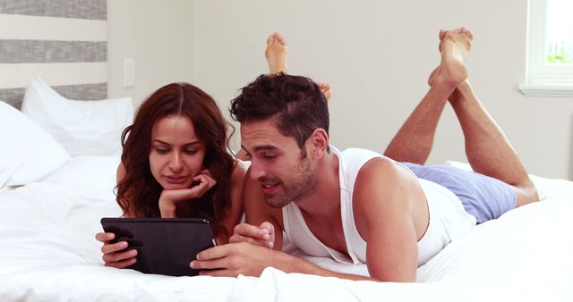 Young couple lying on bed, watching content on tablet together. Great for conveying themes of relaxation, togetherness, modern lifestyles, technology usage, and leisure. Ideal for use in ads, blog posts, or social media promoting technology, relationship goals, and home comfort.