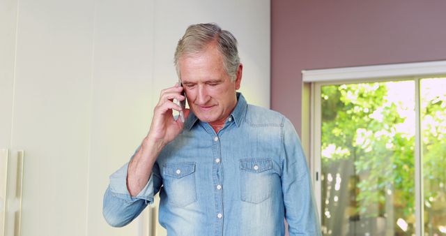 Senior man having a conversation on a smartphone while standing indoors wearing a denim shirt. Ideal for illustrating themes related to elder communication, staying connected with family, technology use among seniors, and contemporary home settings.