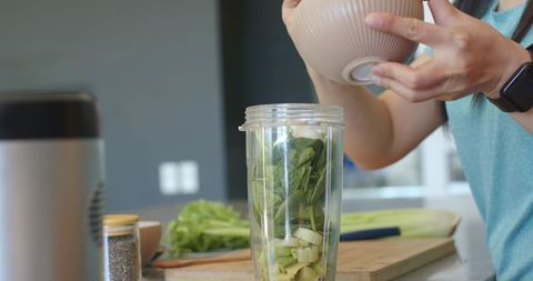 Asian woman pouring green ingredients into blender cup for healthy spinach smoothie