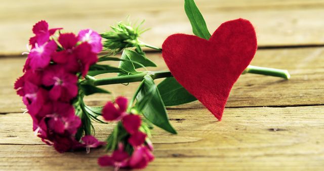 A vibrant bouquet of pink flowers lies next to a red felt heart on a wooden surface, with copy space. These symbols of affection and love are often associated with romantic gestures and special occasions like Valentine's Day.