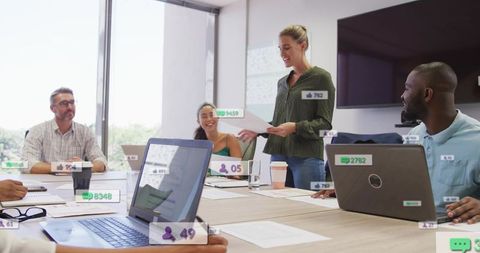 Presenting woman leading collaborative meeting in bright boardroom with laptops and overlays