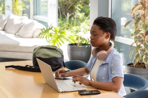 African teenage woman studying on laptop in sunlit lounge with pink headphones and plants