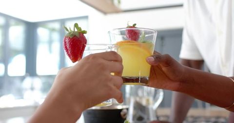 Diverse friends clinking citrus drinks garnished with strawberry and orange in bright kitchen