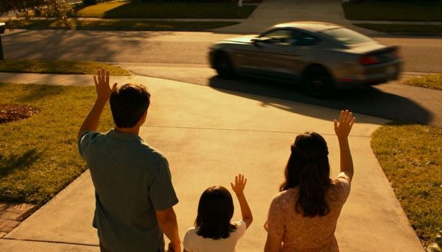 Family waving from driveway at passing car on suburban street during golden hour evening