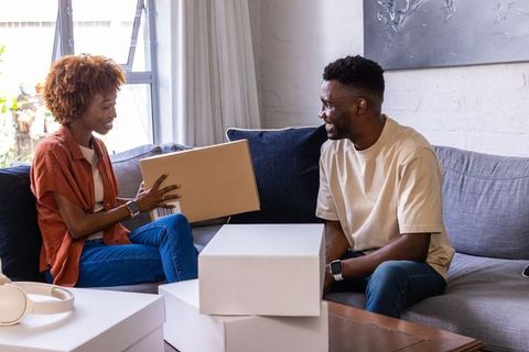 Couple exchanging parcel on sofa, unpacking boxes at home, cozy modern living room lifestyle smiling