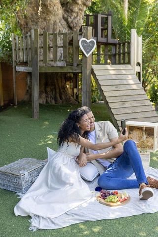 Romantic backyard picnic with diverse couple taking selfie beside treehouse