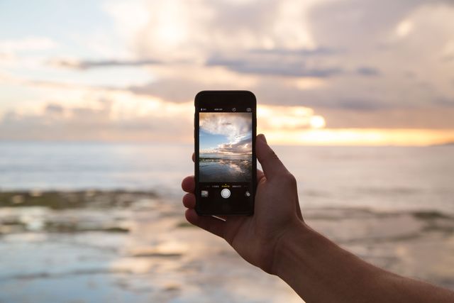 Hand holding smartphone photographing beautiful sunset over ocean, creating serene and tranquil moment ideal for promoting travel, technology, and outdoor activities.